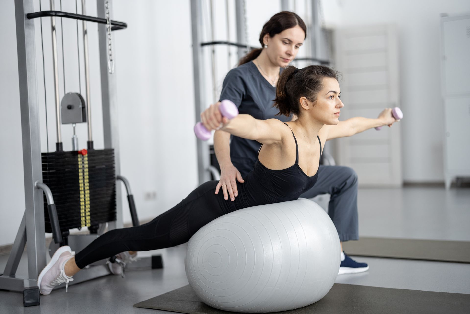 A woman is sitting on a pilates ball while a female physical therapist holds dumbbells.