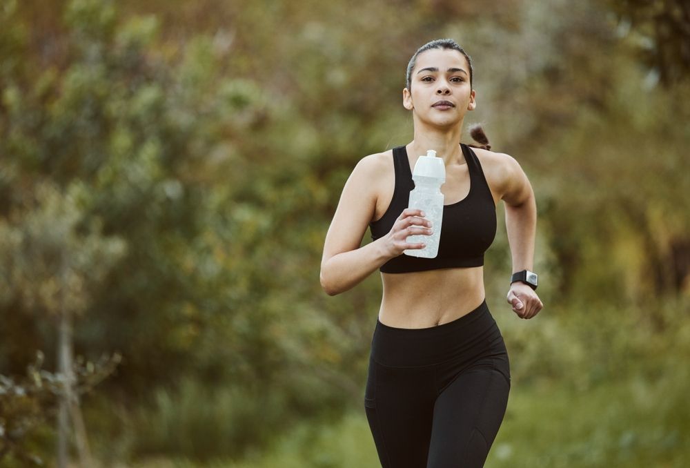 Woman running outdoors in black athletic wear, holding a water bottle in a blurred natural setting