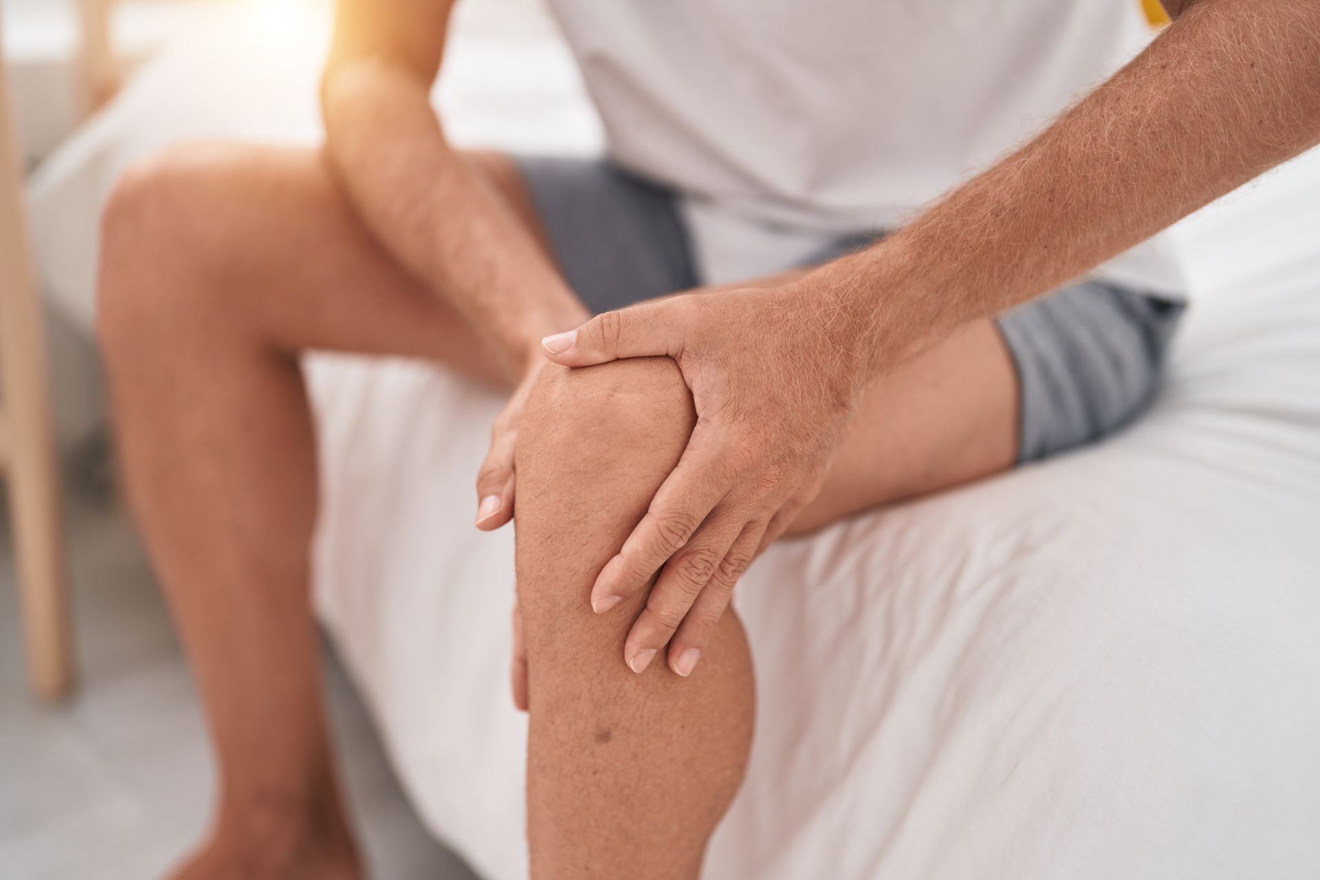 A man is sitting on a bed holding his knee in pain.