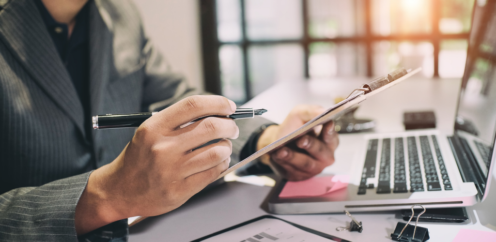 Person in a suit writing on a clipboard, laptop and office supplies on the desk.