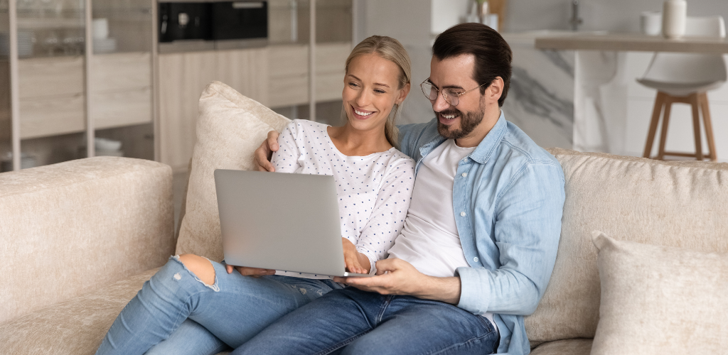 Couple sitting on a couch, looking at a laptop together, smiling. Modern living room setting.