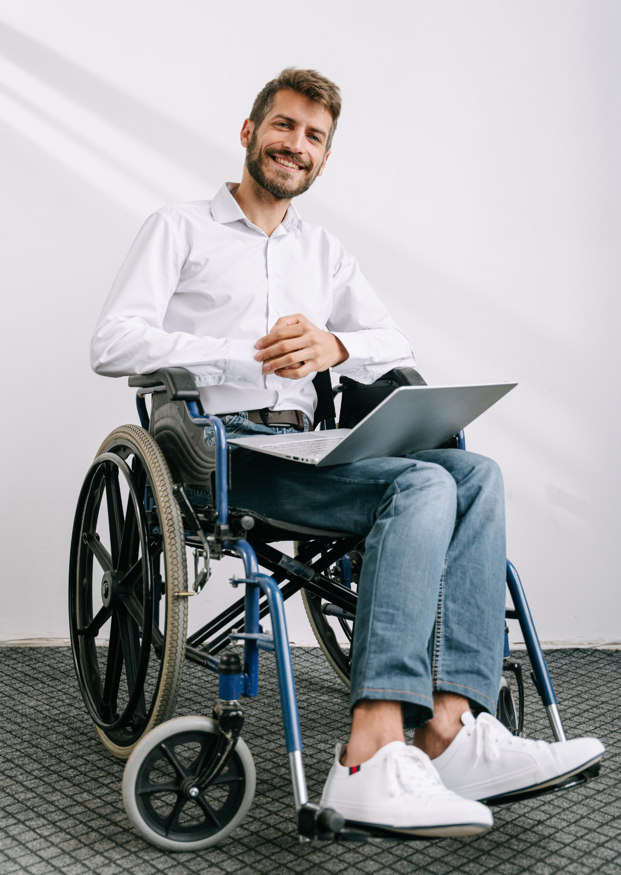 Man in wheelchair smiling, holding laptop, wearing white shirt and jeans. Against a white wall.
