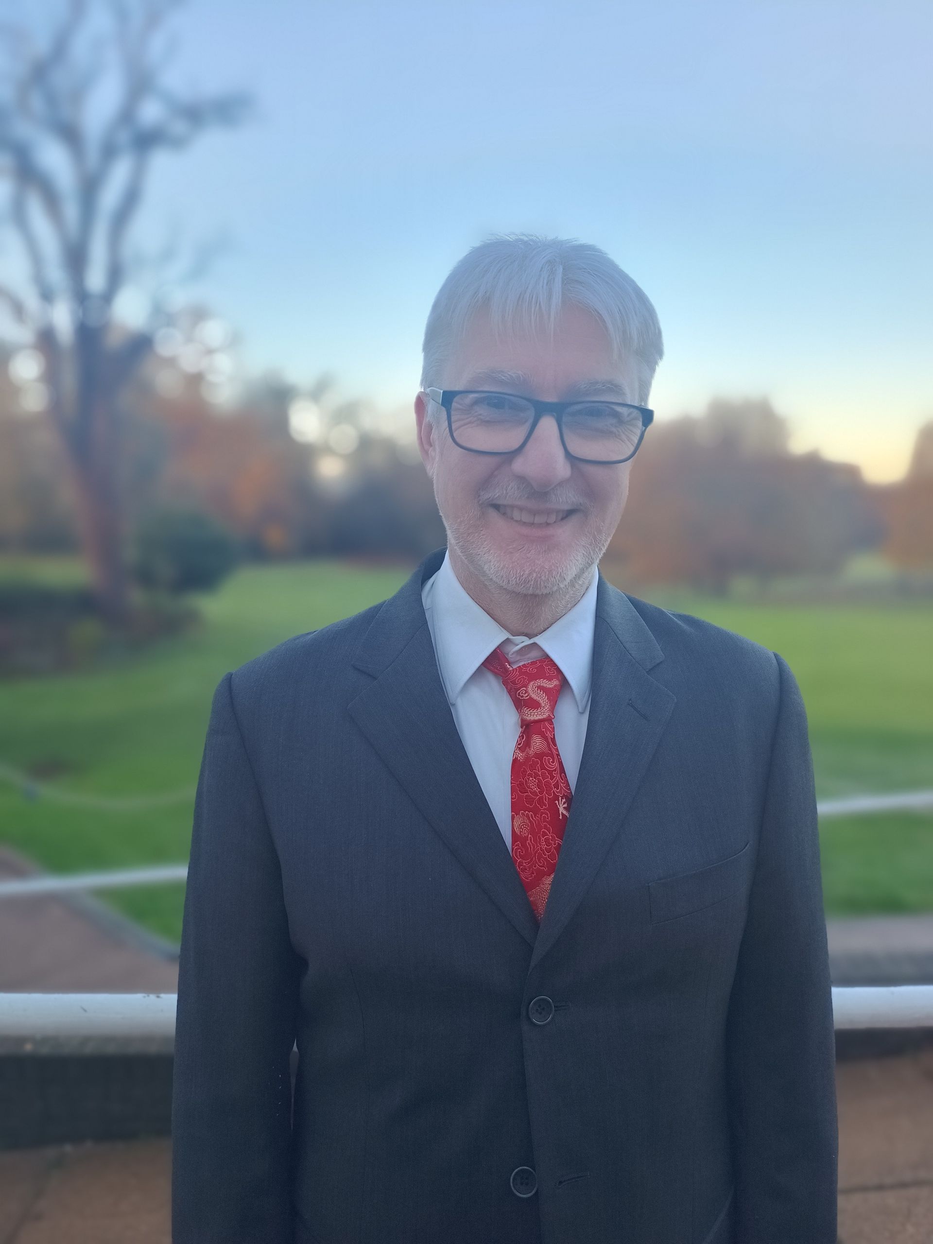 Man in a suit, tie, and glasses smiling outdoors with park scenery in background.