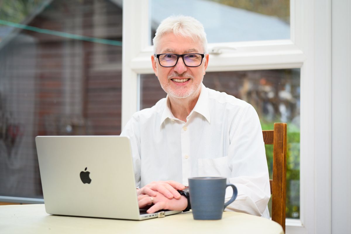 Man in a suit, tie, and glasses smiling outdoors with park scenery in background.