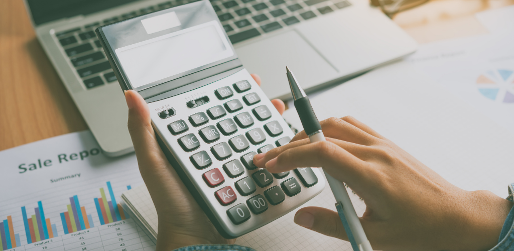 Person using a calculator with a pen, laptop, and sales report visible.