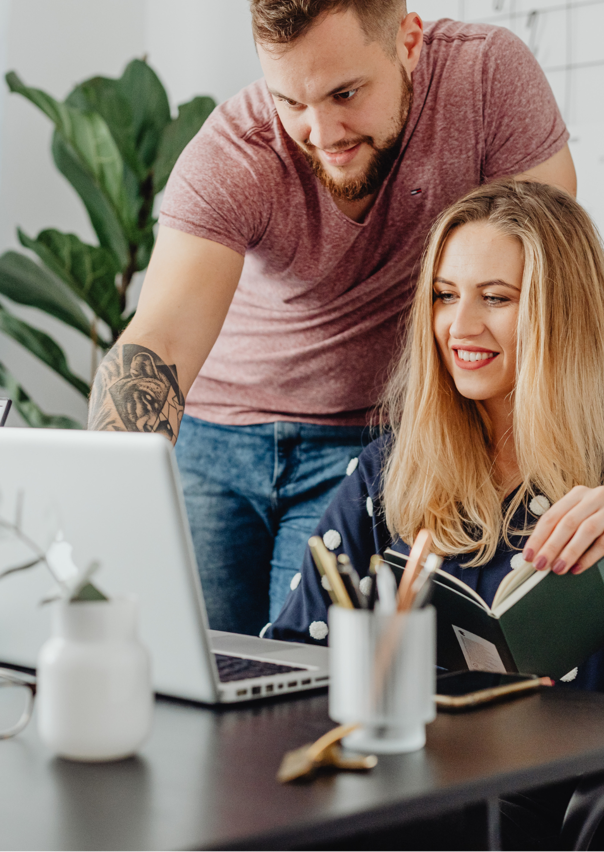 Man and woman looking at a laptop, woman holding notebook, man with arm tattoo, office setting.
