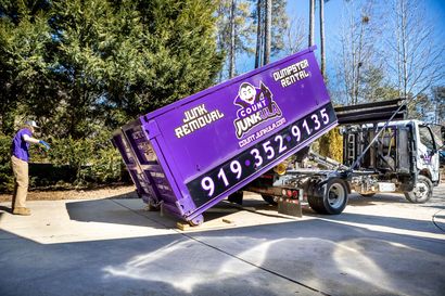 A purple junk removal dumpster being loaded onto a truck by a man in a blue shirt.