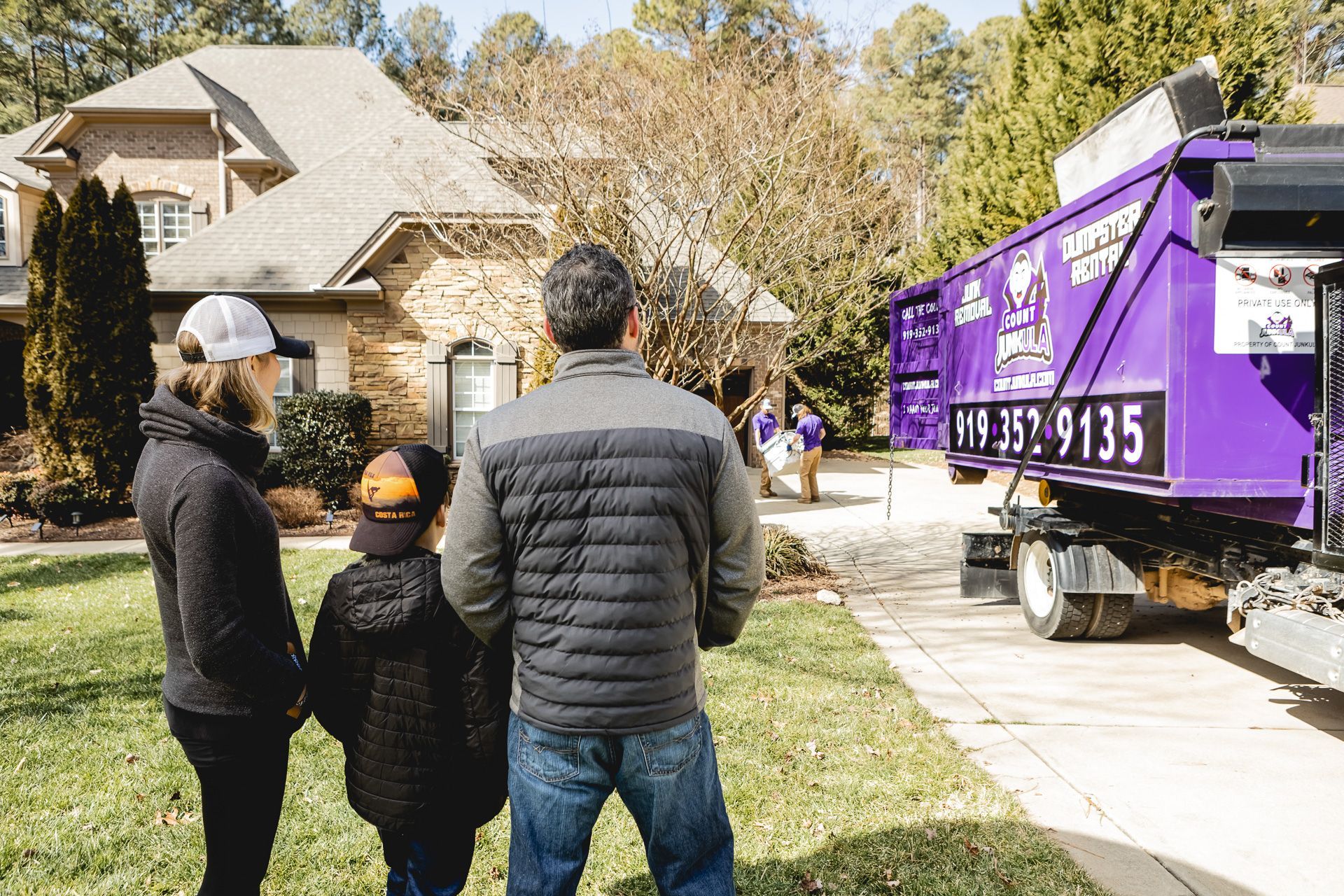 Family watches a purple dumpster being delivered to their house on a sunny day