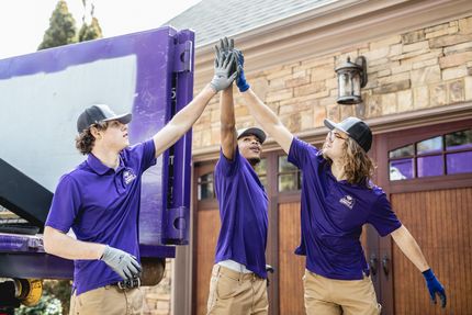 Three people in purple shirts high-fiving in front of a truck, near a house with a stone exterior.