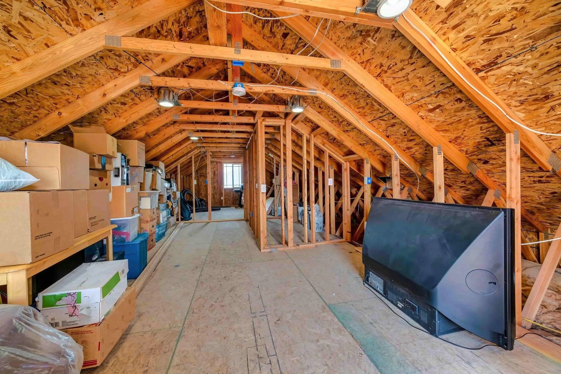 Attic storage space with exposed wooden beams, plywood floor, and stacked boxes. A large TV rests against the wall.
