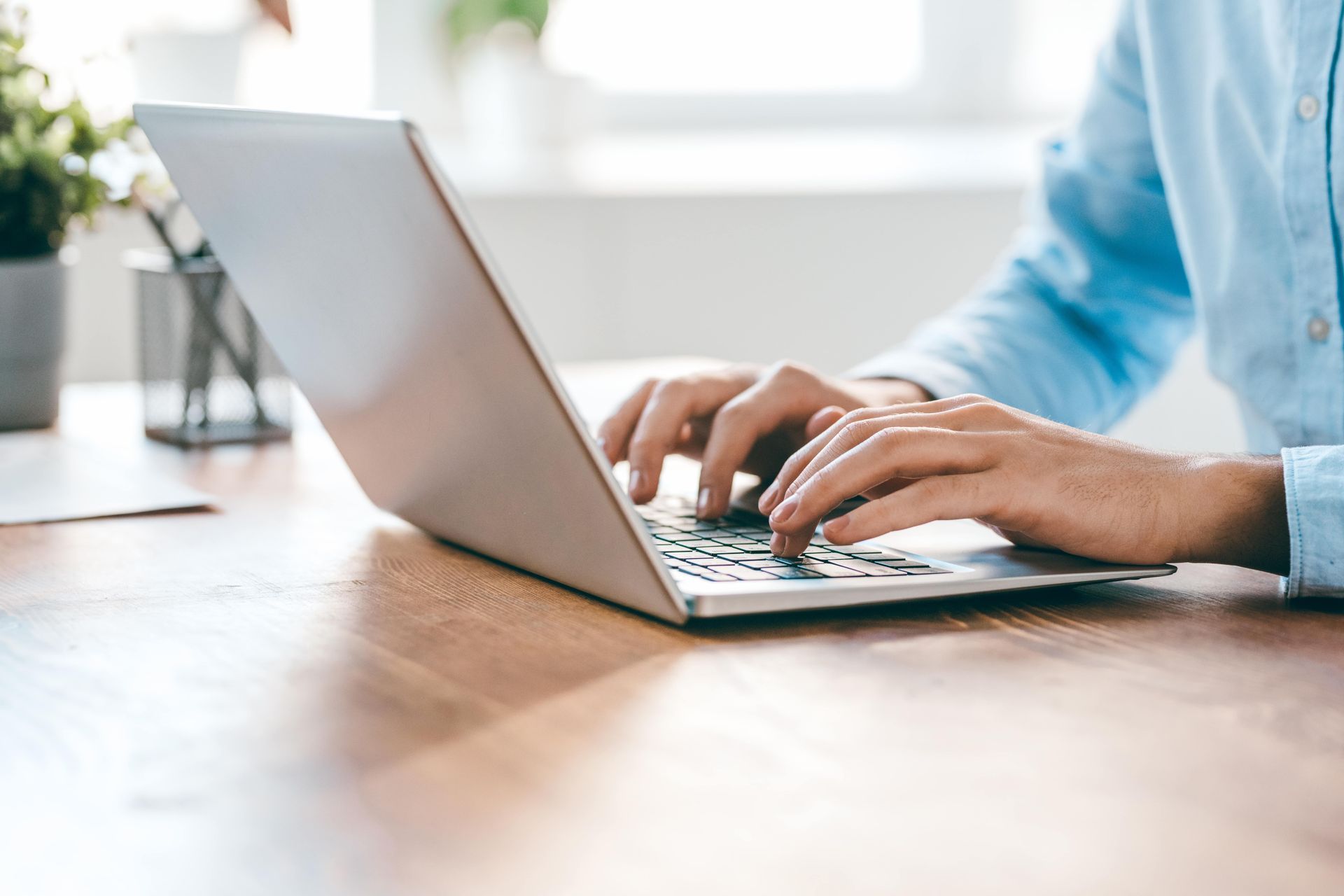 Person typing on a laptop at a wooden desk, wearing a blue shirt.
