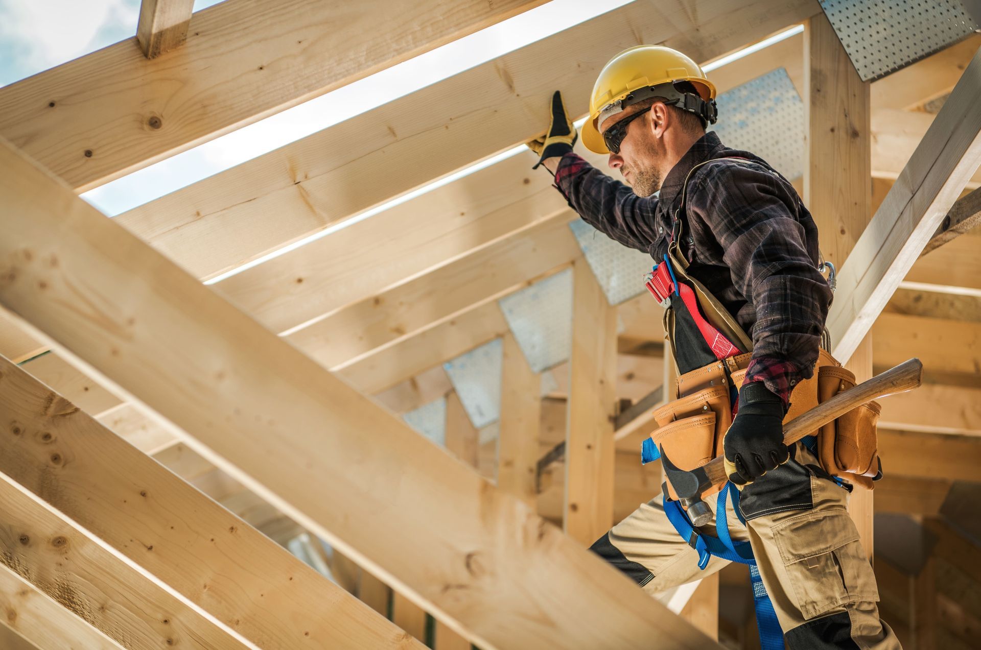 Carpenter in yellow hard hat, safety harness, and tool belt working on wooden roof frame.
