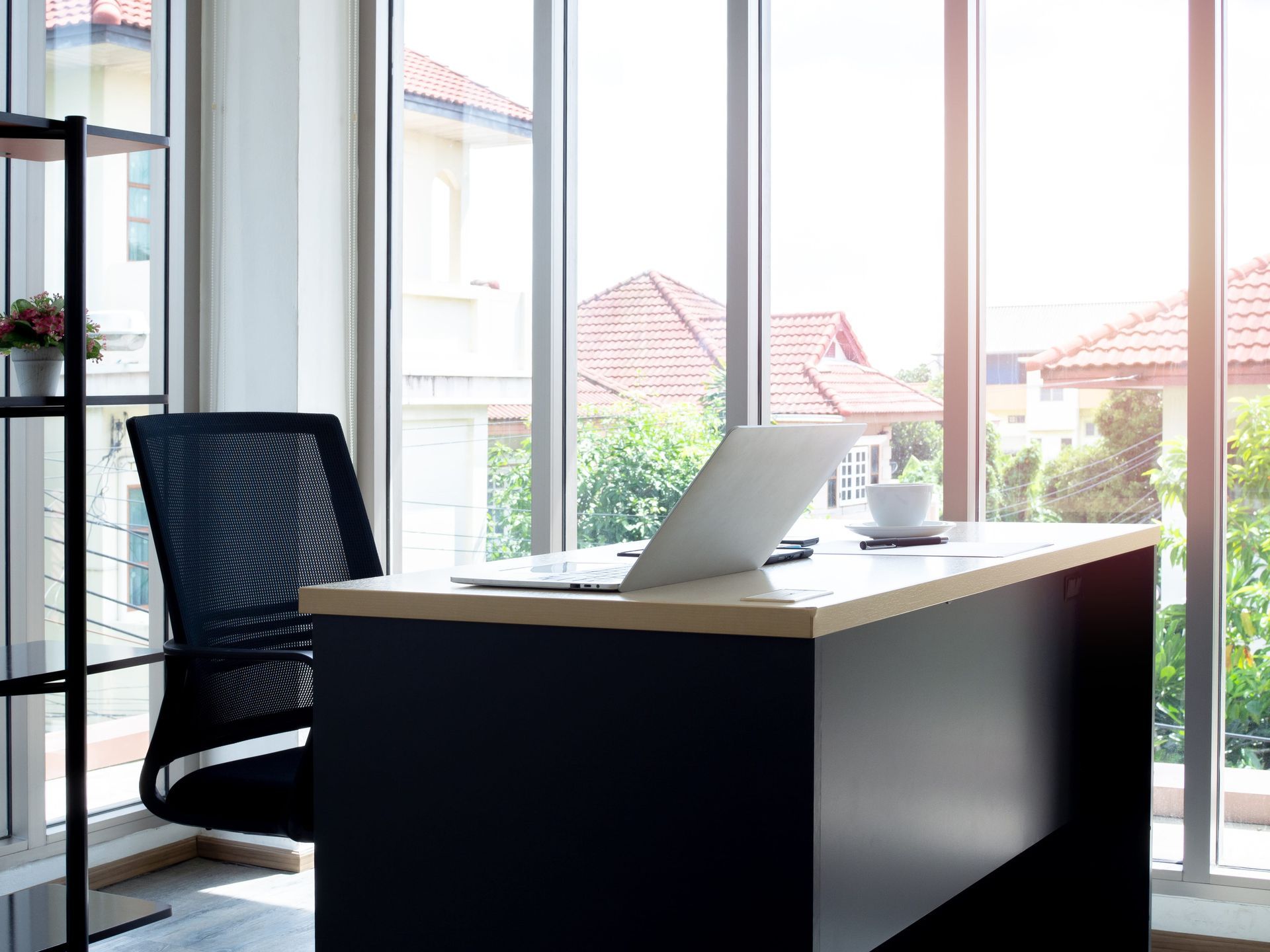 Office workspace with desk, laptop, chair, and large window overlooking houses.