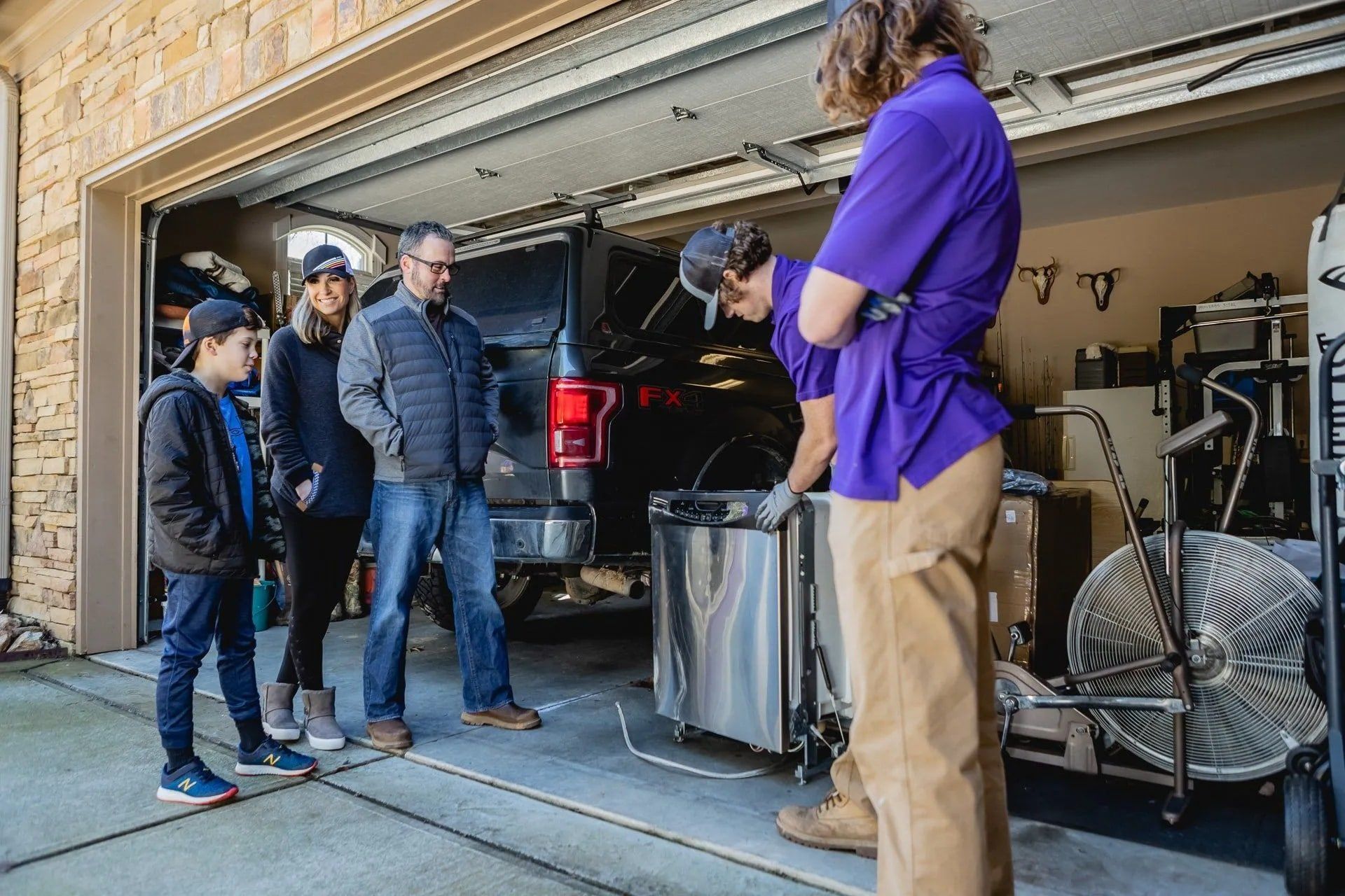 Family watching a repair person in a purple shirt work on an appliance in a garage.