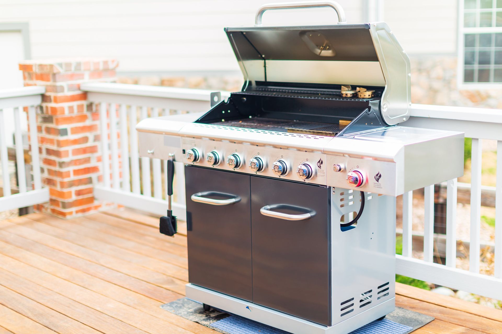 Gas grill on a wooden deck, lid open. Stainless steel with six burner controls.