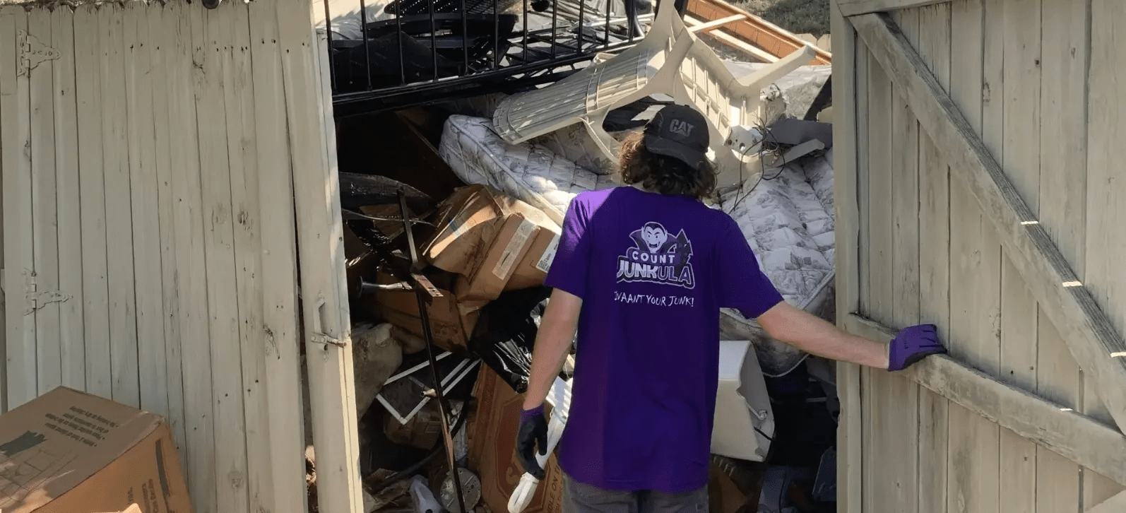 A person in purple shirt opens a shed door filled with debris, another person is inside the shed.