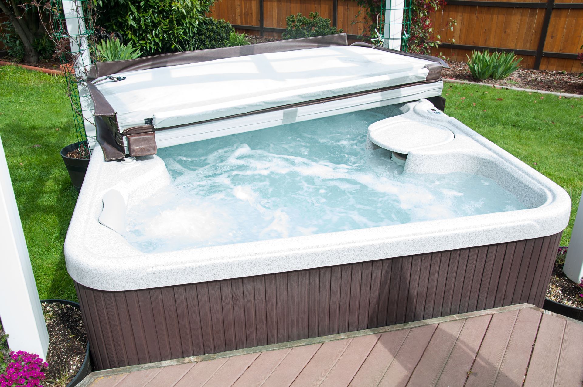 A bubbling hot tub in a backyard, brown siding and cover, on a small wooden deck, green grass.