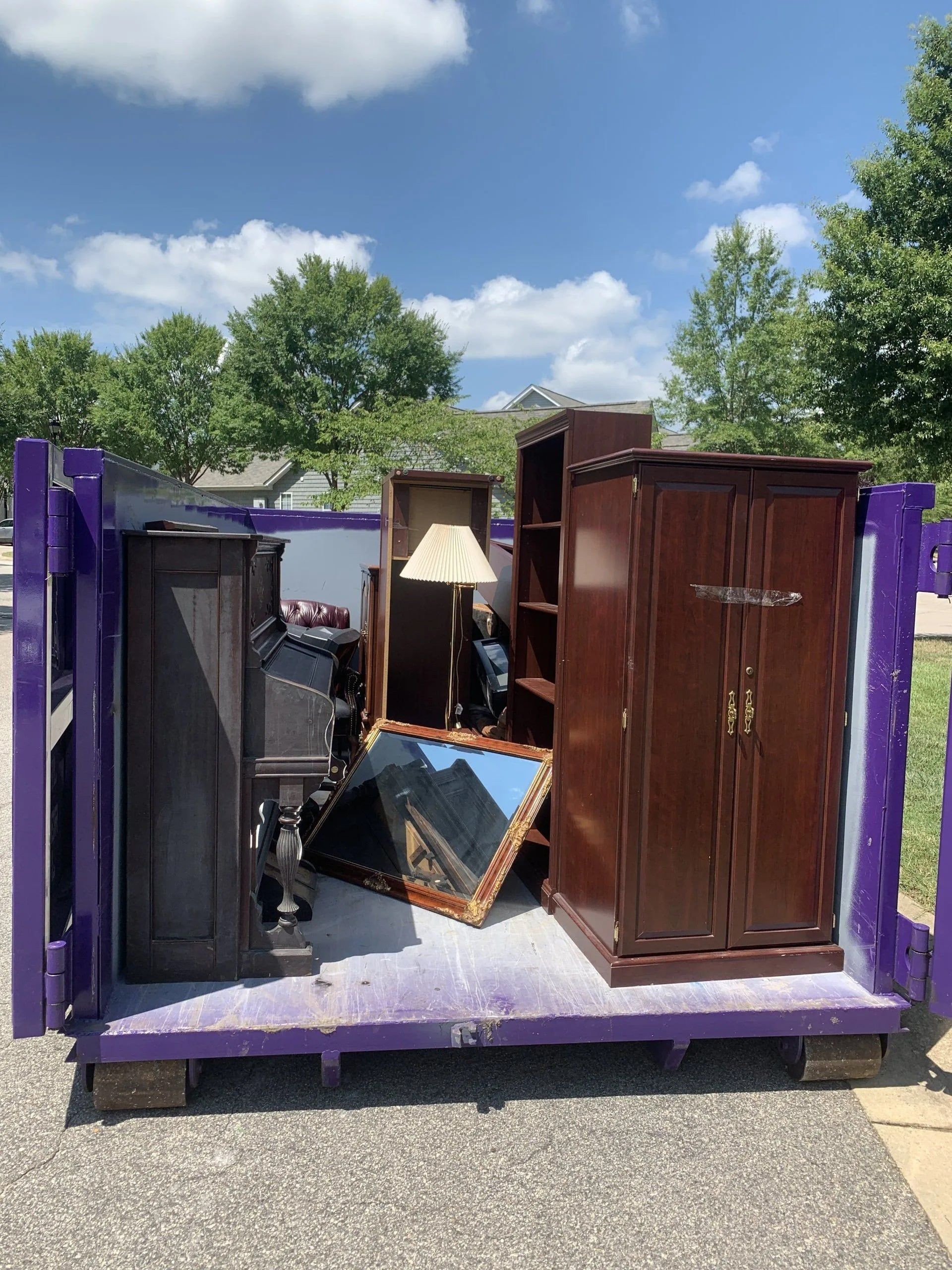 A purple dumpster filled with discarded furniture: a wardrobe, shelves, lamp, and a mirror, under a blue sky.