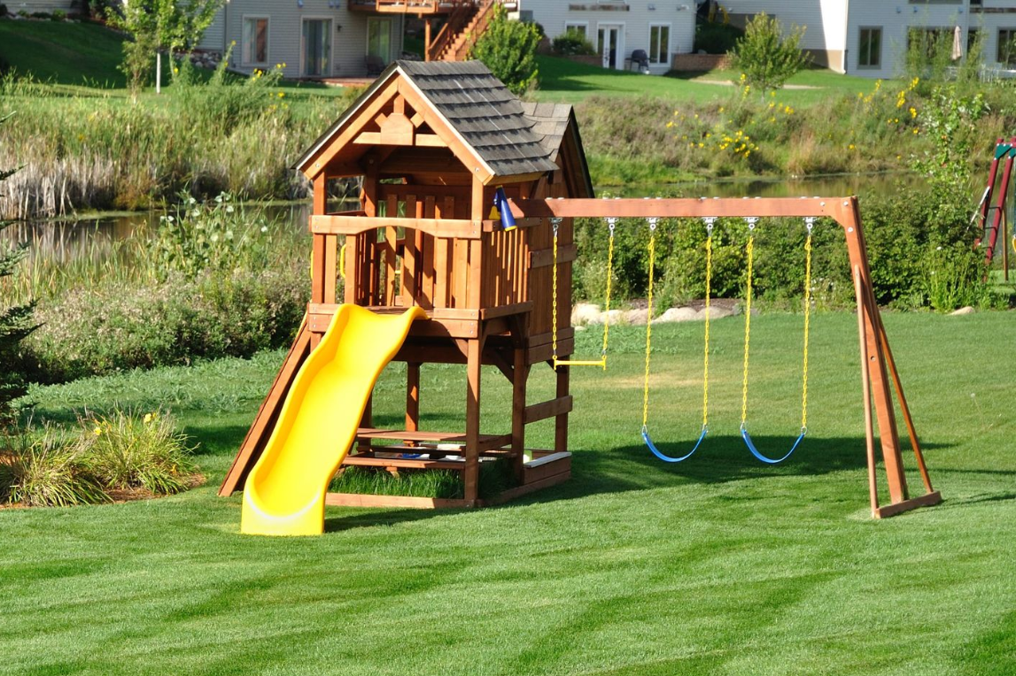 Wooden playset with a yellow slide and swings on a green lawn.