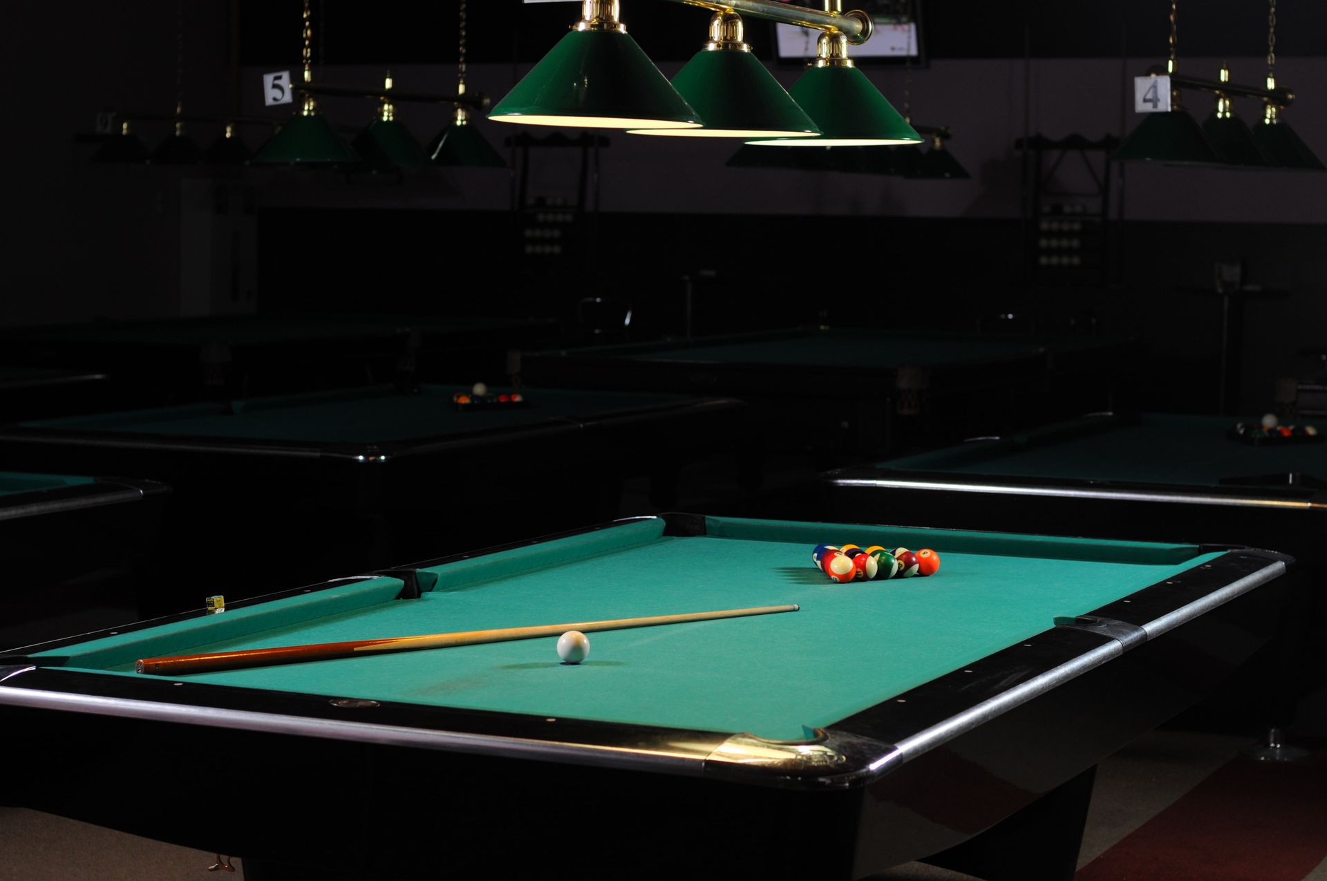 A dimly lit pool hall with several green felt tables, overhead lamps, and a rack of billiard balls.