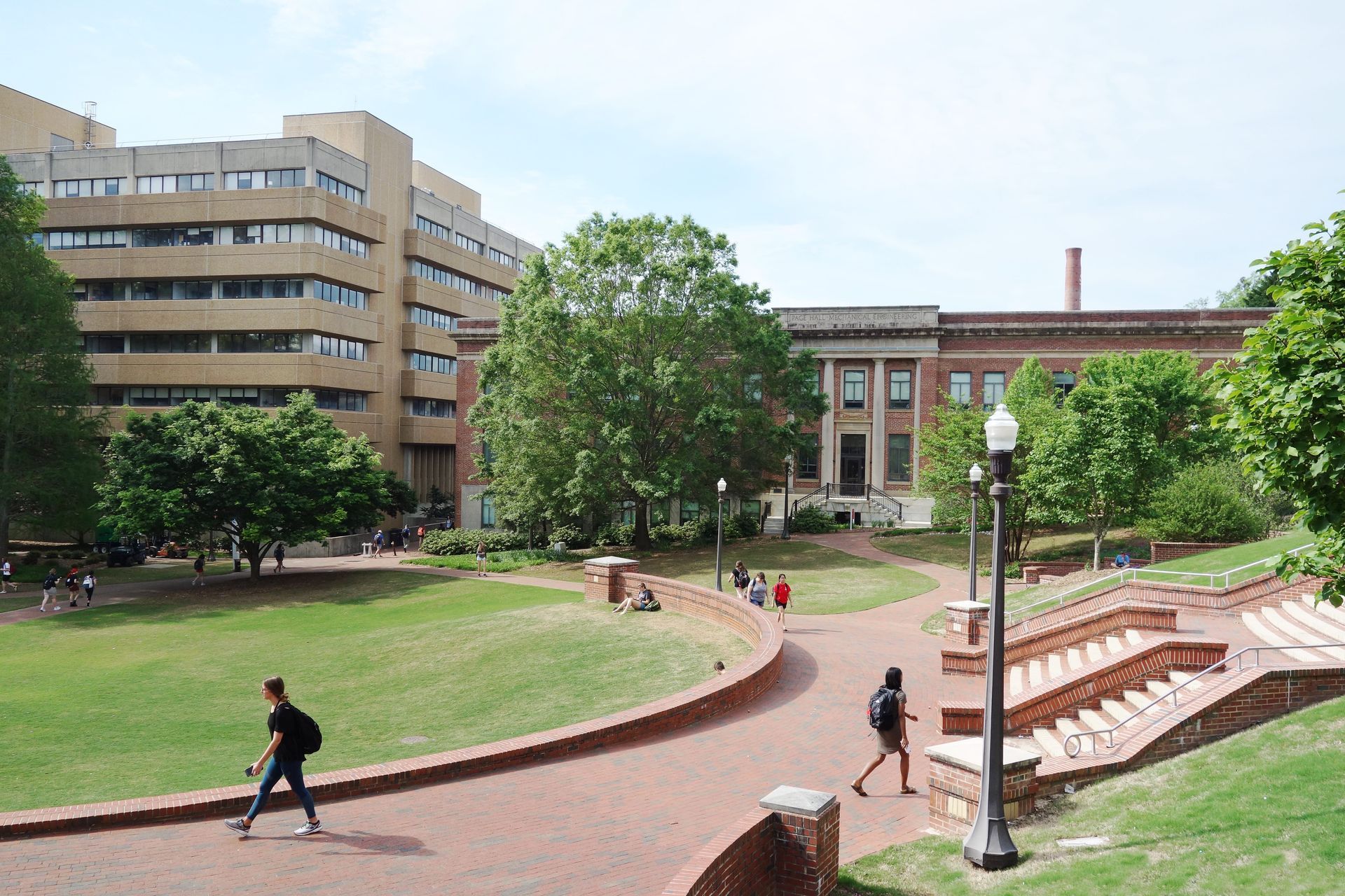 Brick path through a green campus with two people walking. Buildings and trees surround. Sunny day.