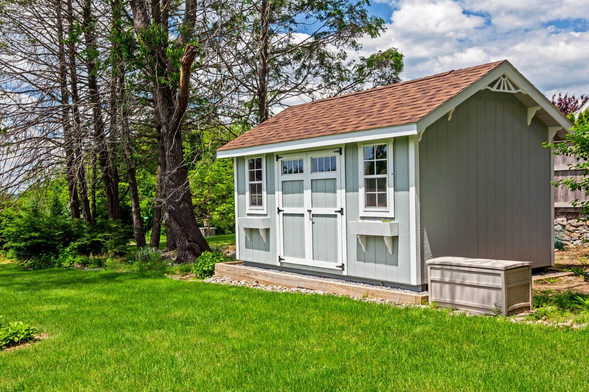 Gray shed with white-trimmed doors and windows, brown roof, set on green grass.