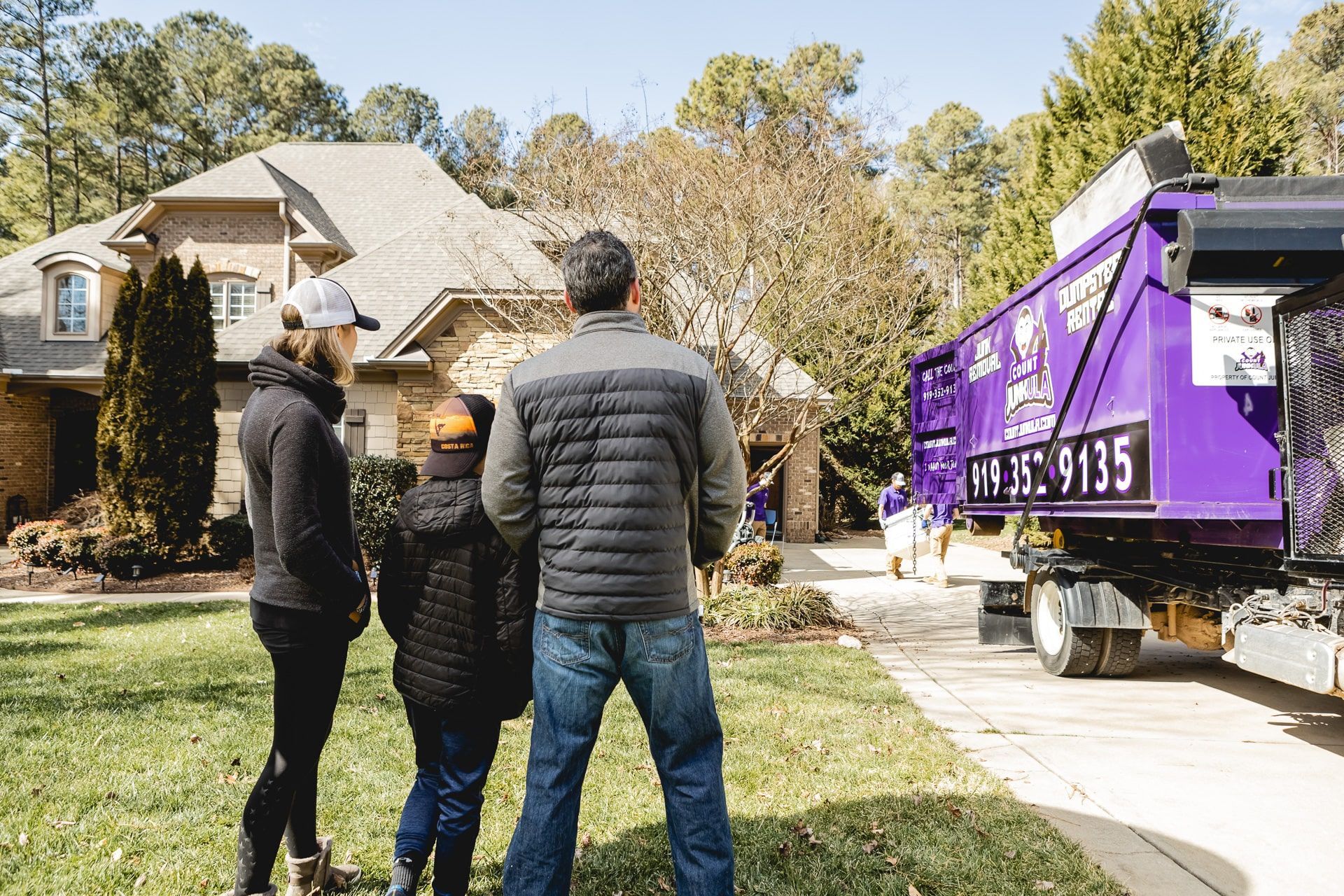 Family watching a purple dumpster truck in front of a house.