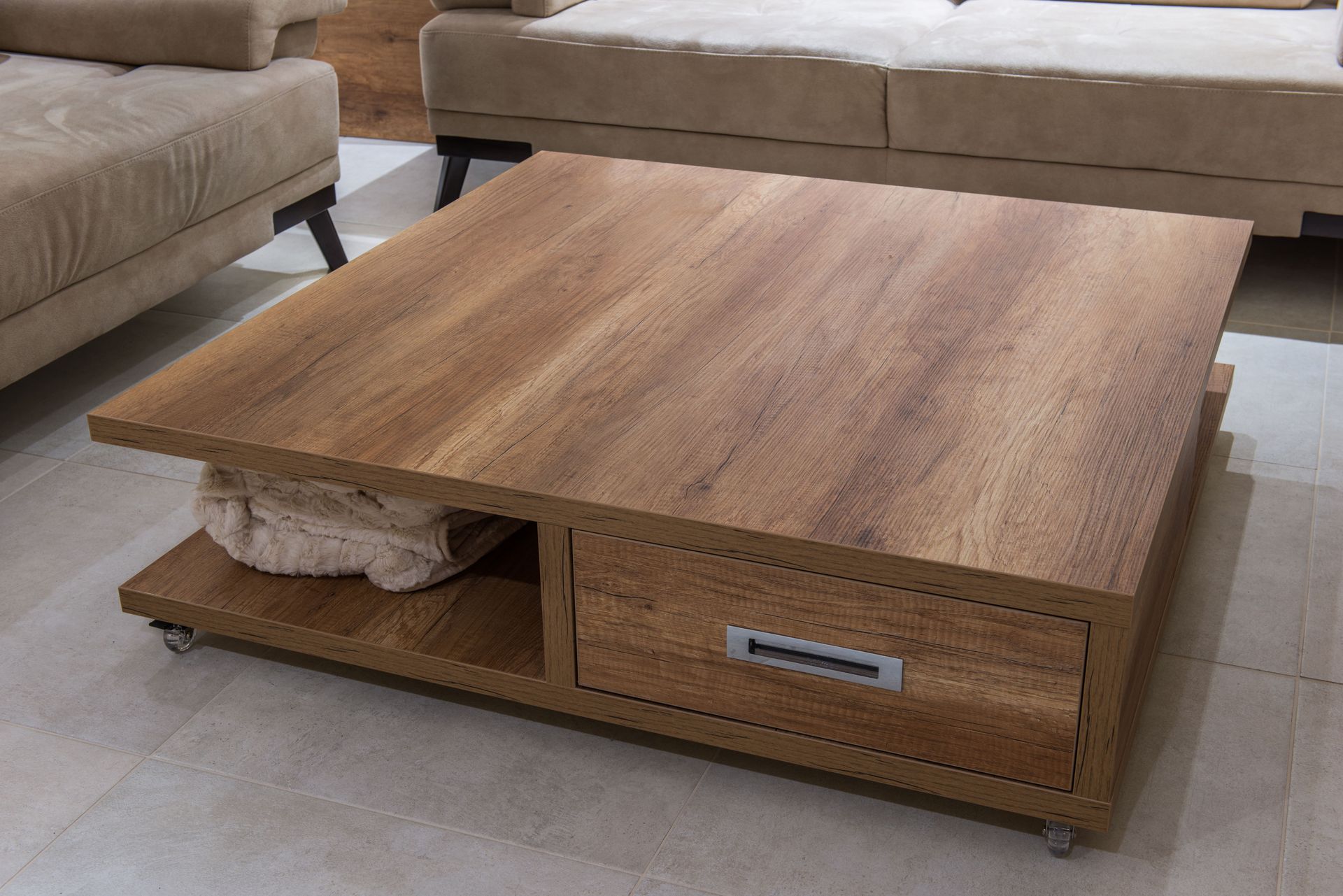 Wooden square coffee table with drawer, lower shelf, and casters, in front of a beige sofa.