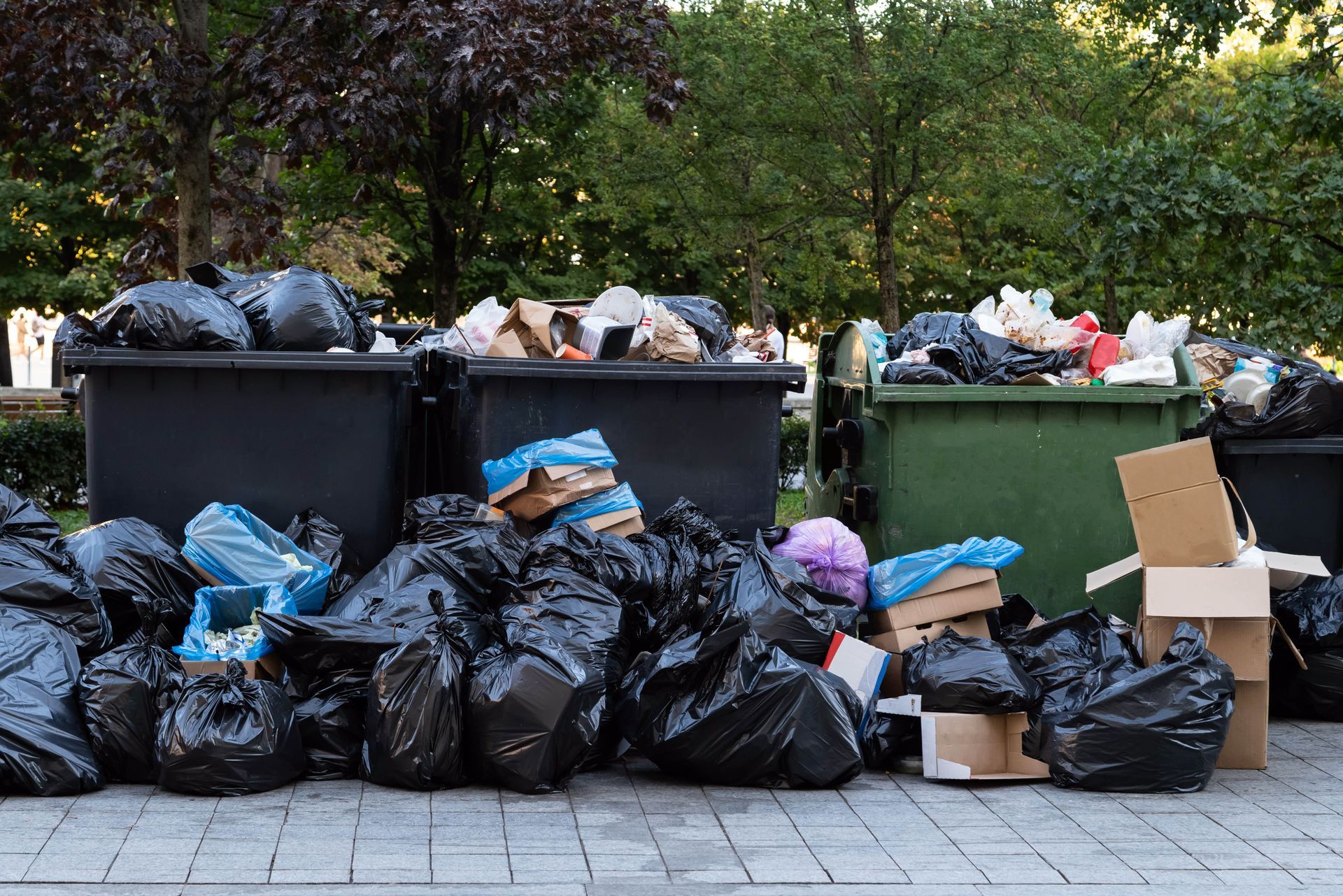 Black garbage bags overflowing beside two dumpsters. Boxes and trash litter the area outdoors.