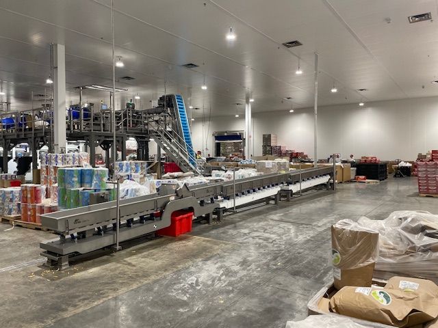 Industrial food processing facility, conveying equipment with packaging. Bright interior, products stacked, long conveyor belt.