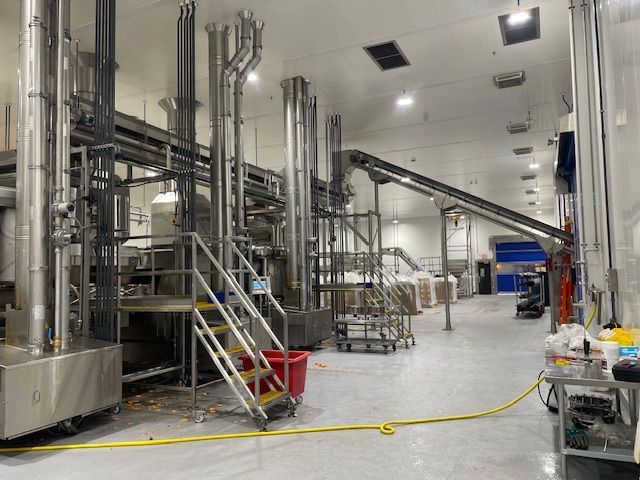 Industrial food processing facility interior with stainless steel pipes, machinery, conveyor belt, and a staircase.