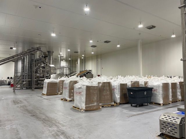 Large, bright industrial food processing facility with pallets of bagged product, machinery, and concrete floor.