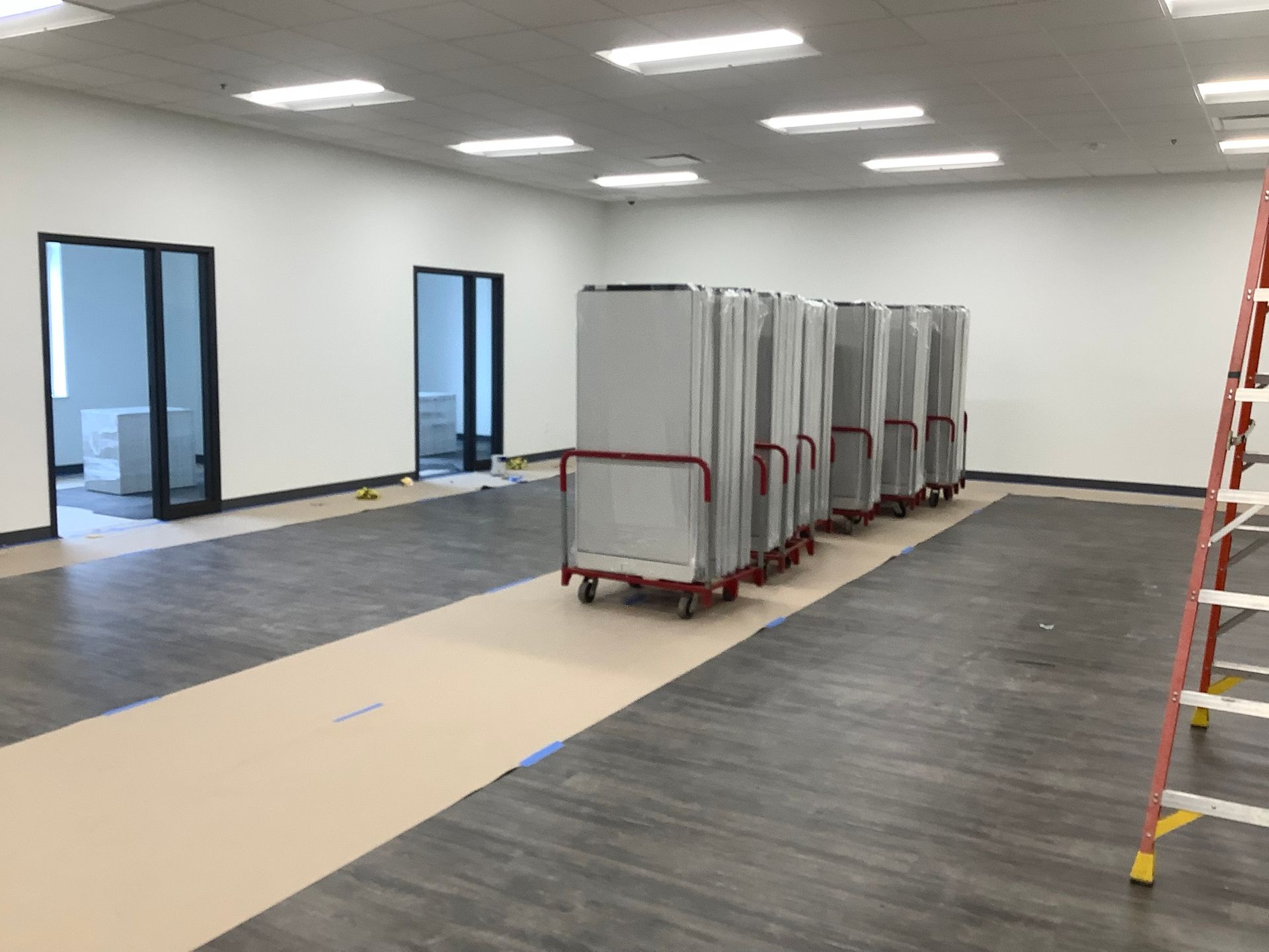 A long row of silver doors on red carts in a nearly empty room with gray flooring, two open black doors, and a ladder.