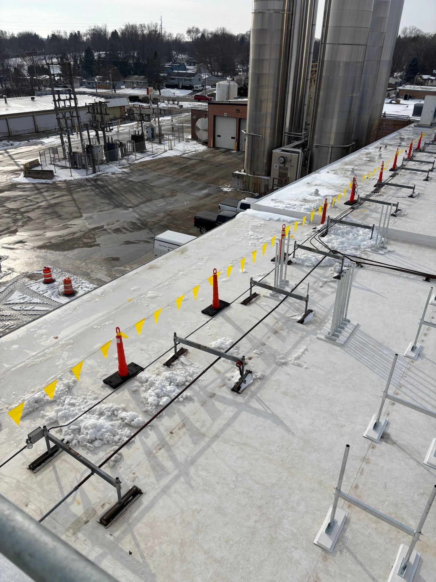 Rooftop with safety barriers: a row of supports, yellow flags, and traffic cones, near silos and a snowy industrial area.