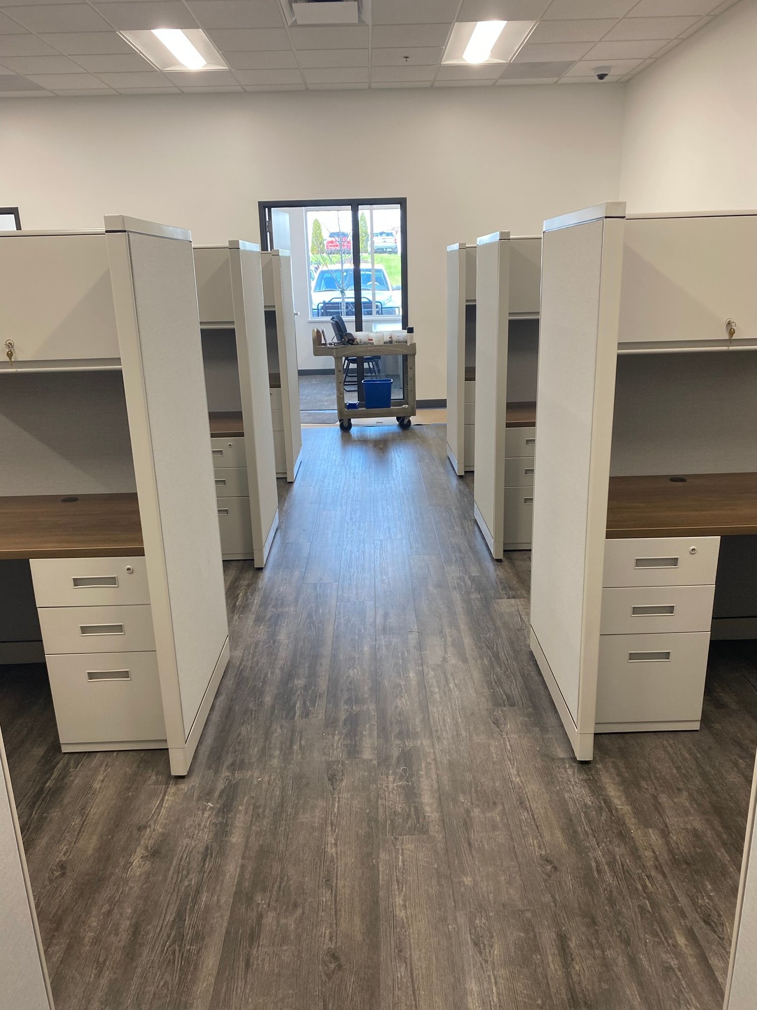 Office cubicles in a row, with white dividers, wood-toned desktops, and gray flooring.