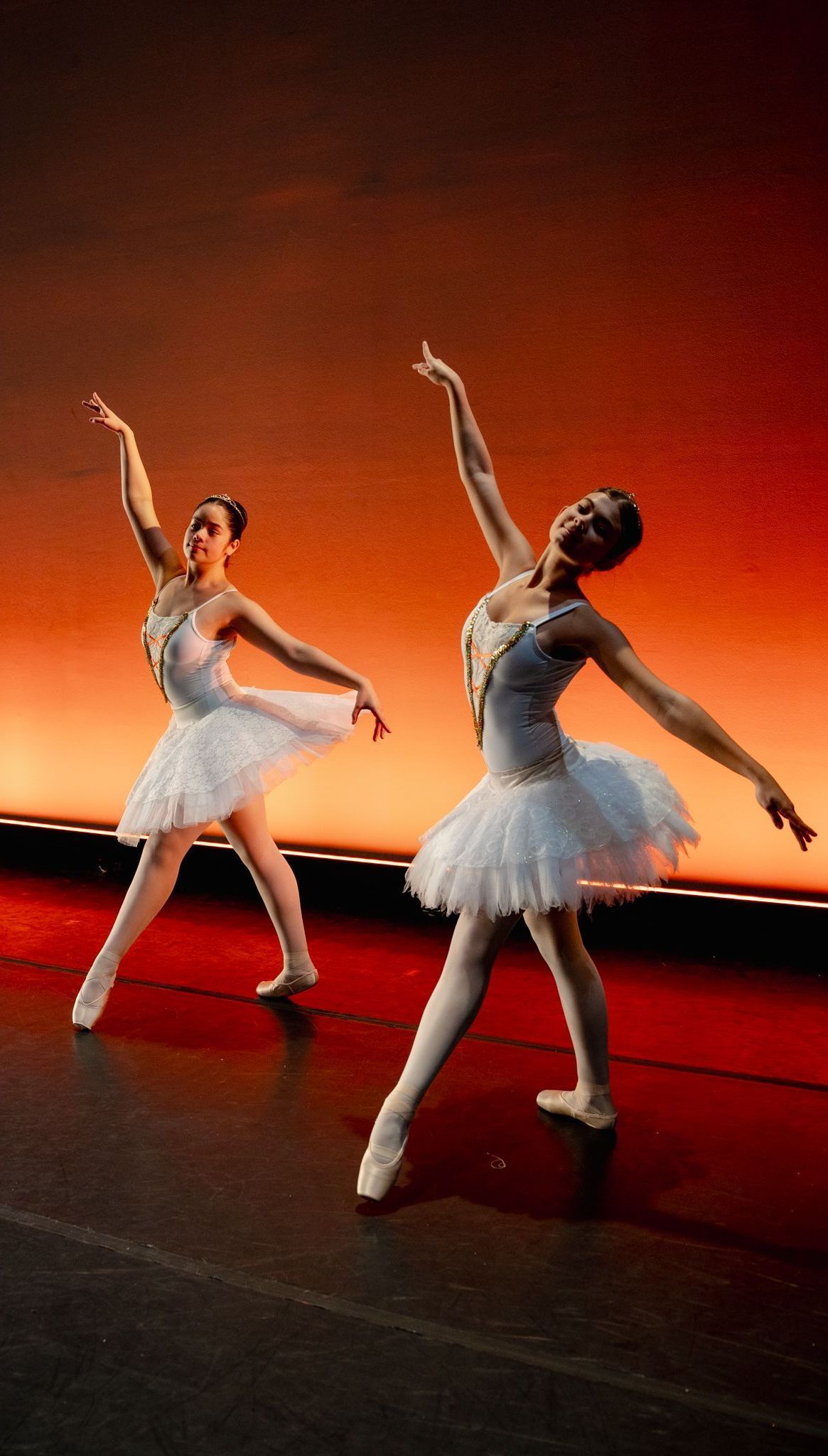 Two ballerinas are dancing on a stage in front of a red background.