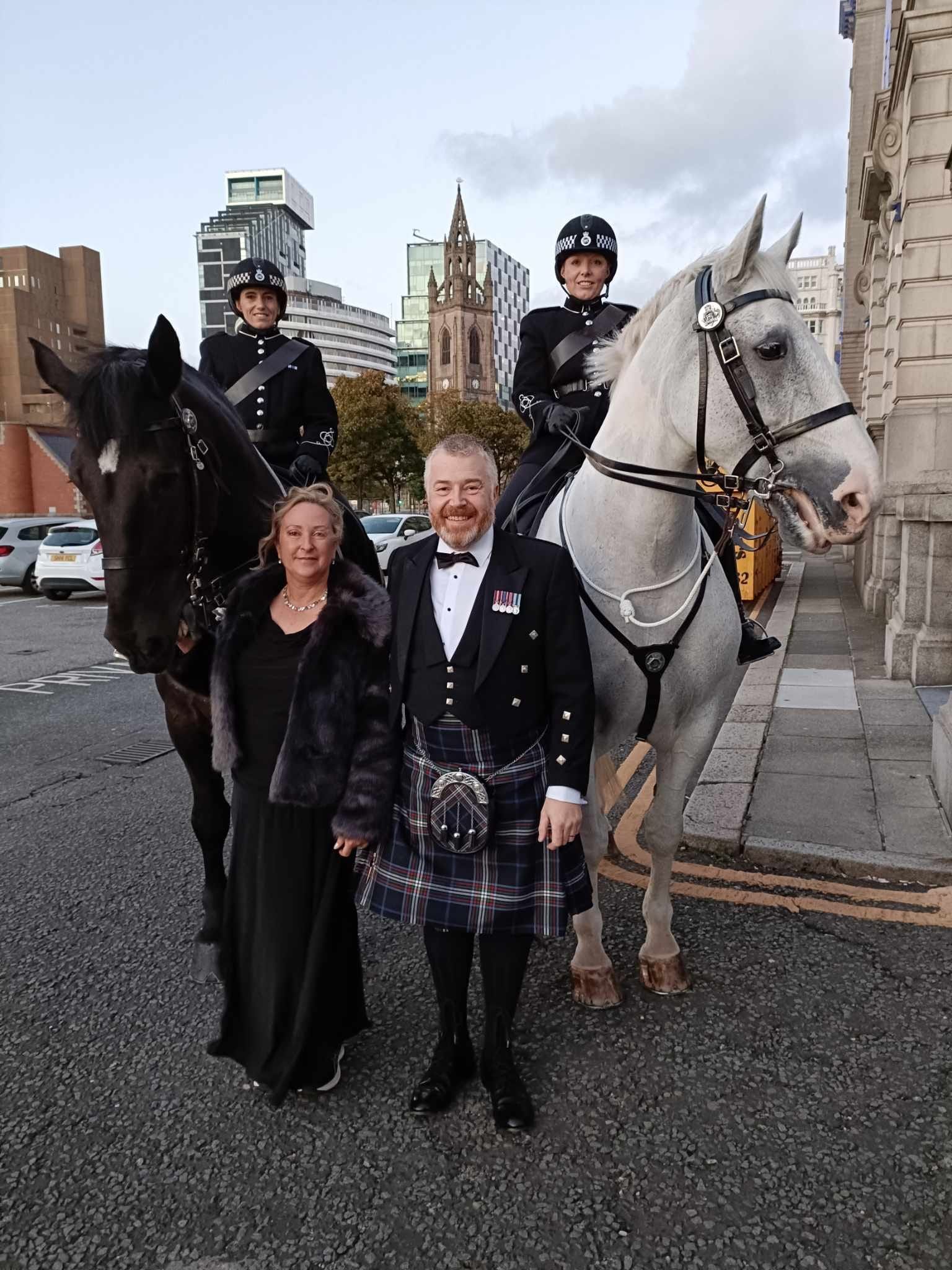 Man in kilt and woman in gown pose with mounted police in Liverpool.