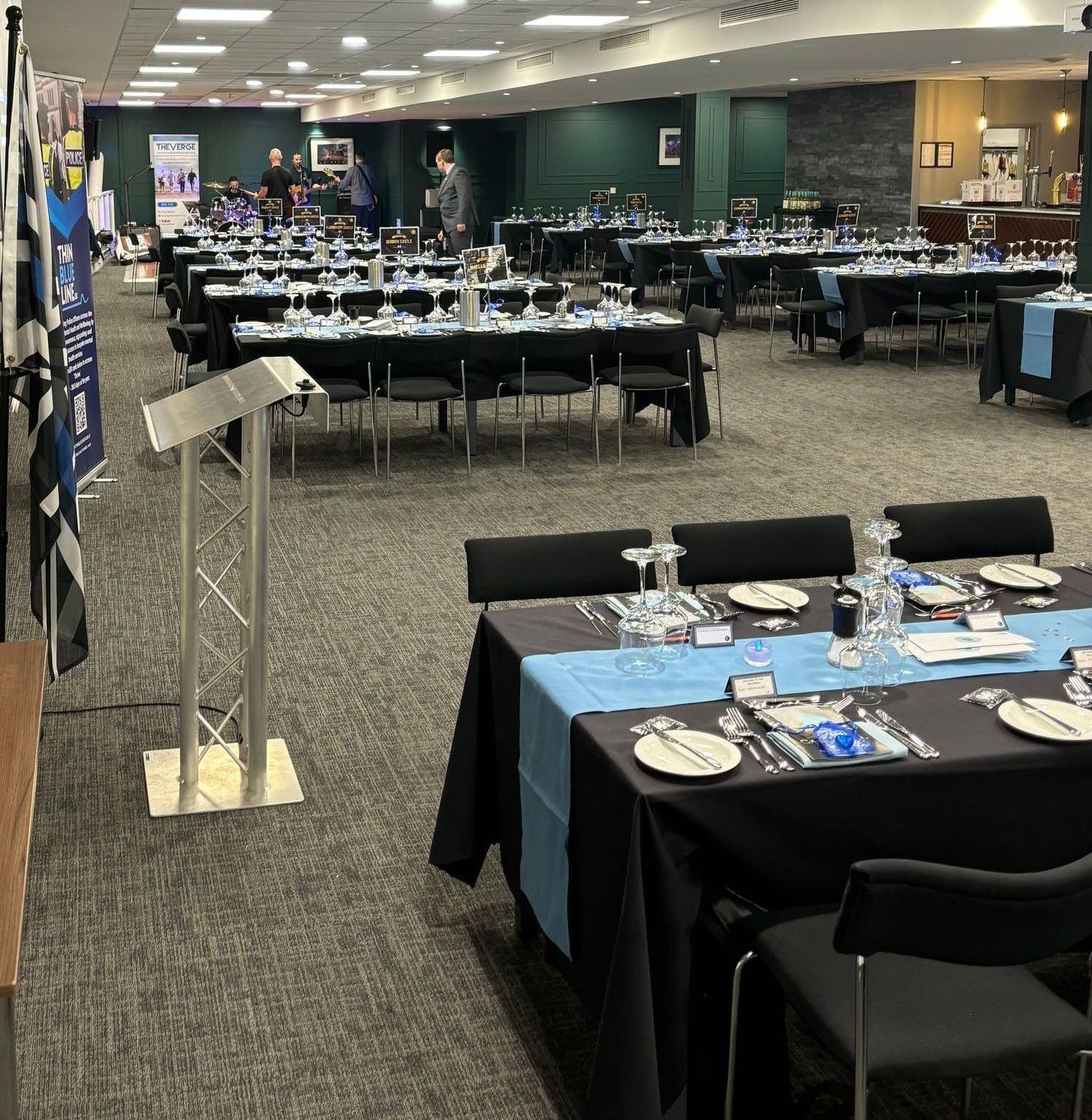 A large event room with tables set for a formal dinner. Black and blue table decor, a podium, and some people are visible.