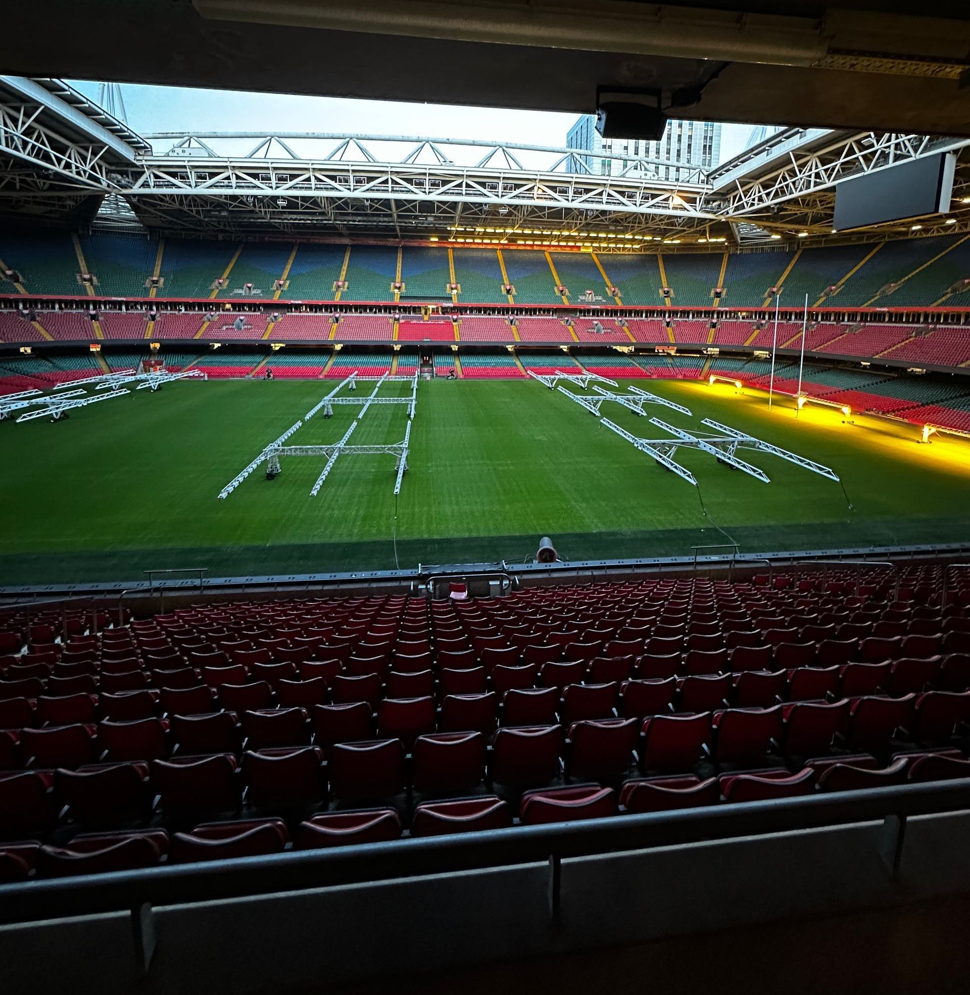 Empty stadium; rows of red seats face a green field with white markings. Bright lights illuminate the scene.