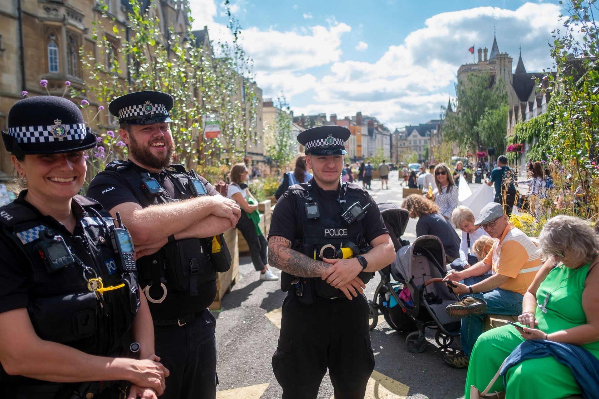 Three smiling police officers standing on a pedestrian street, people seated nearby in the background.