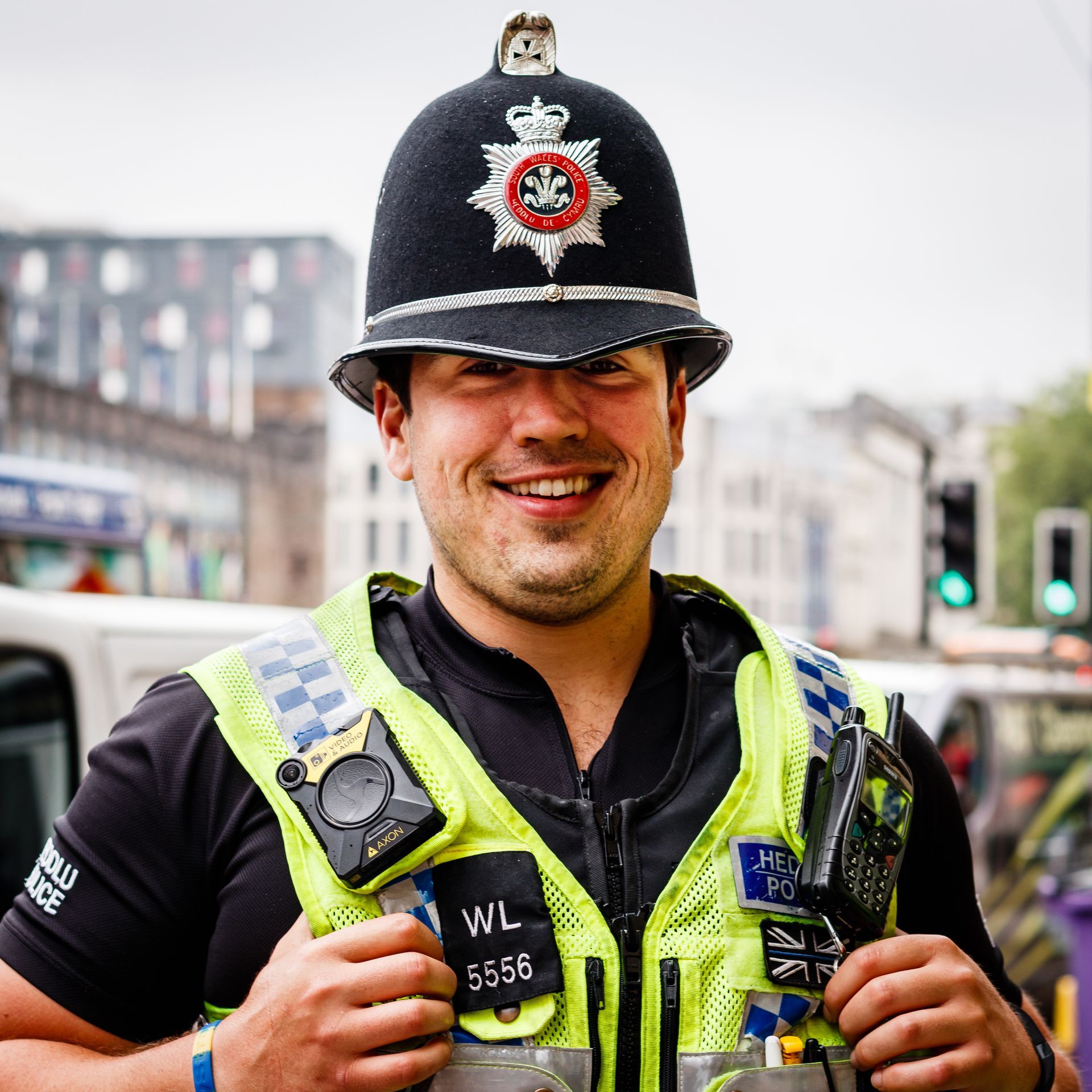 British police officer smiling, wearing a vest, helmet, and uniform in a city.