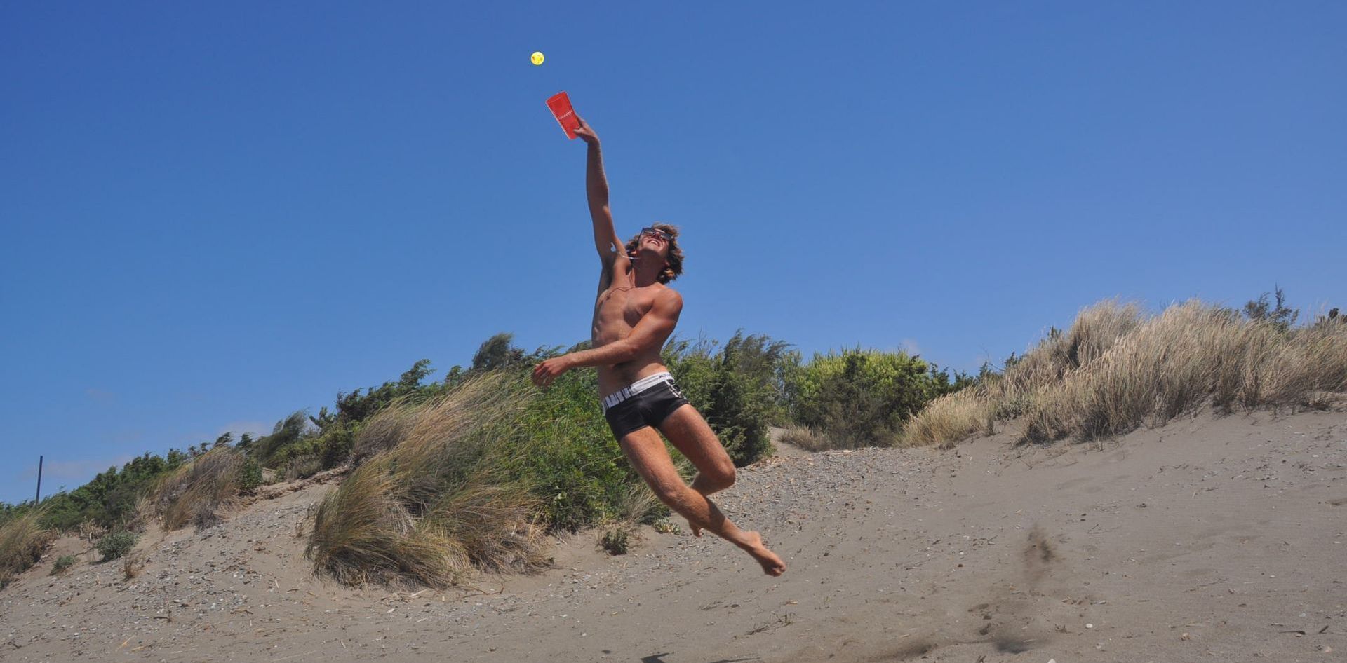 A man leaps to hit a ball with a paddle on a sandy beach under a clear blue sky.