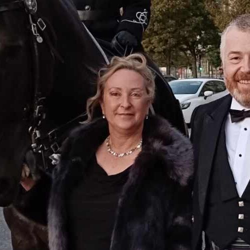 Woman in fur coat smiles, with man in tuxedo. A horse and car are behind them.