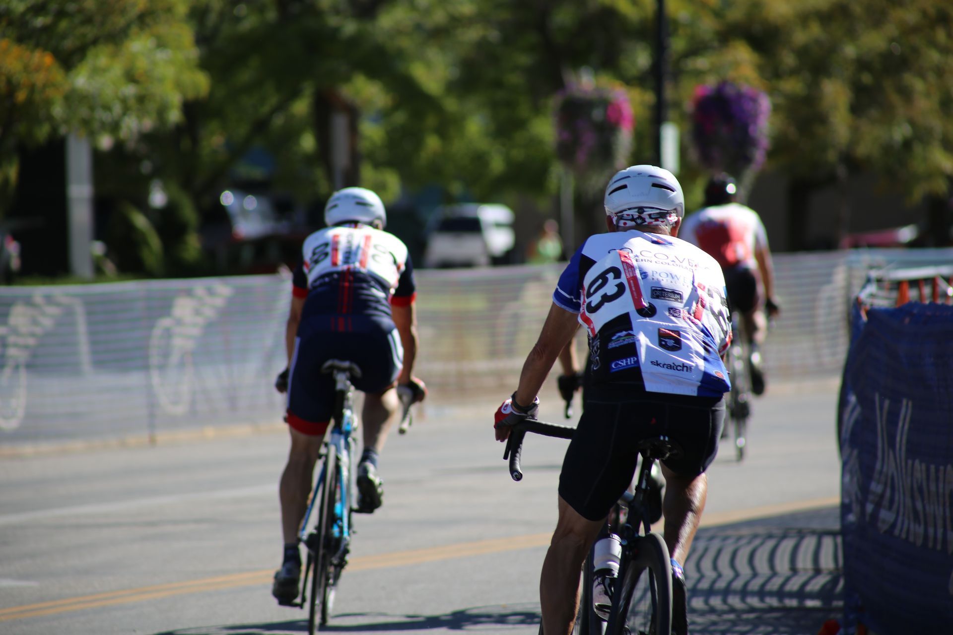 Cyclists in colorful jerseys race on a sunny road, with a barrier and hanging flowers nearby.