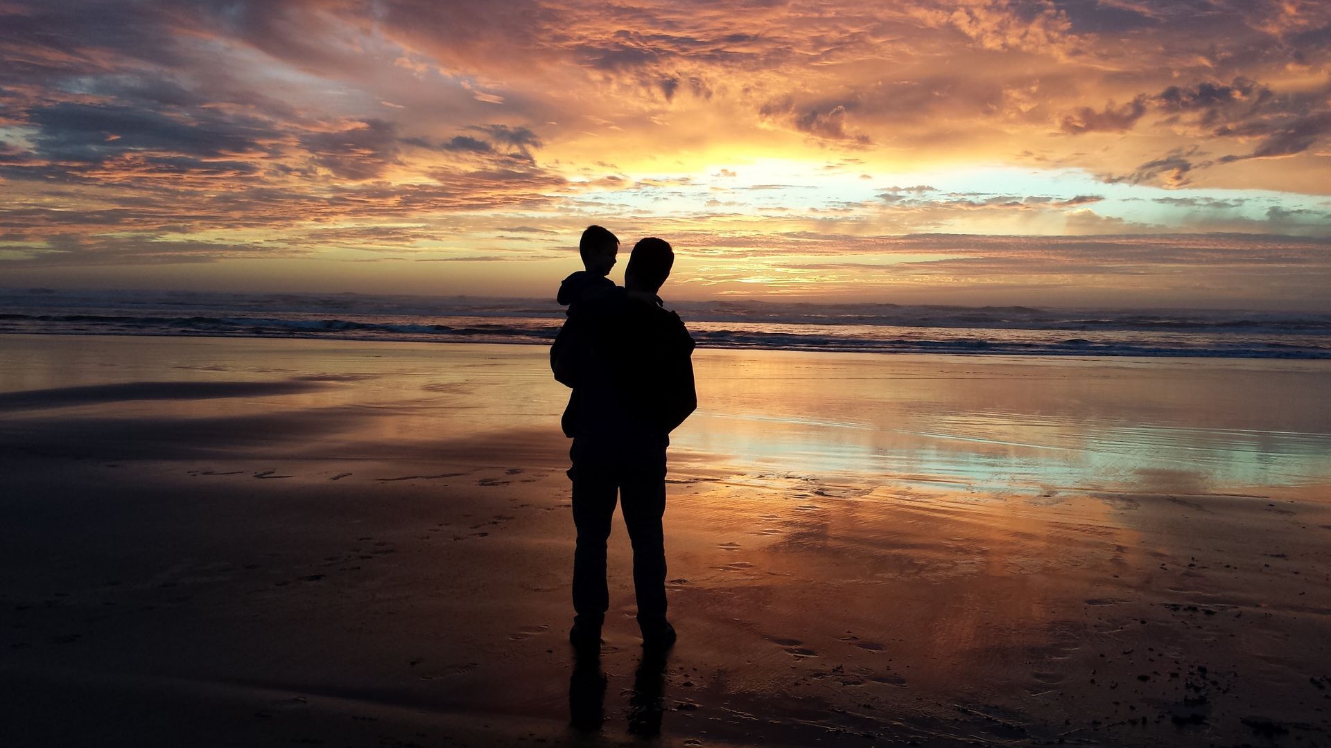 Silhouette of a father carrying a child on a beach, watching a colorful sunset over the ocean.