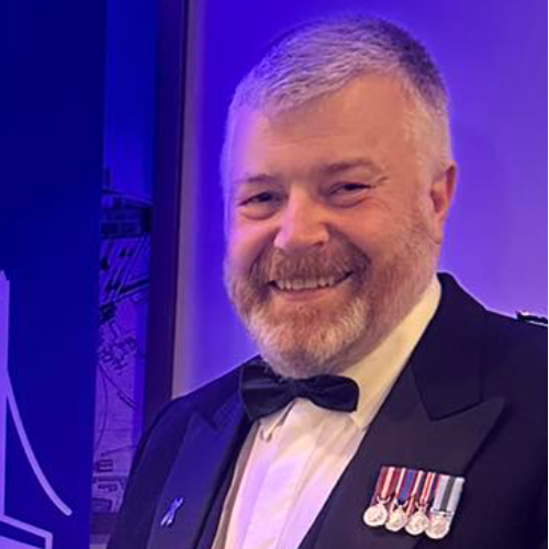 Man in formalwear, smiling, with medals, against a blue backdrop.