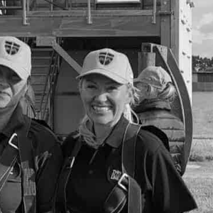 Woman smiling, wearing cap, harness, and black shirt, with others in background.