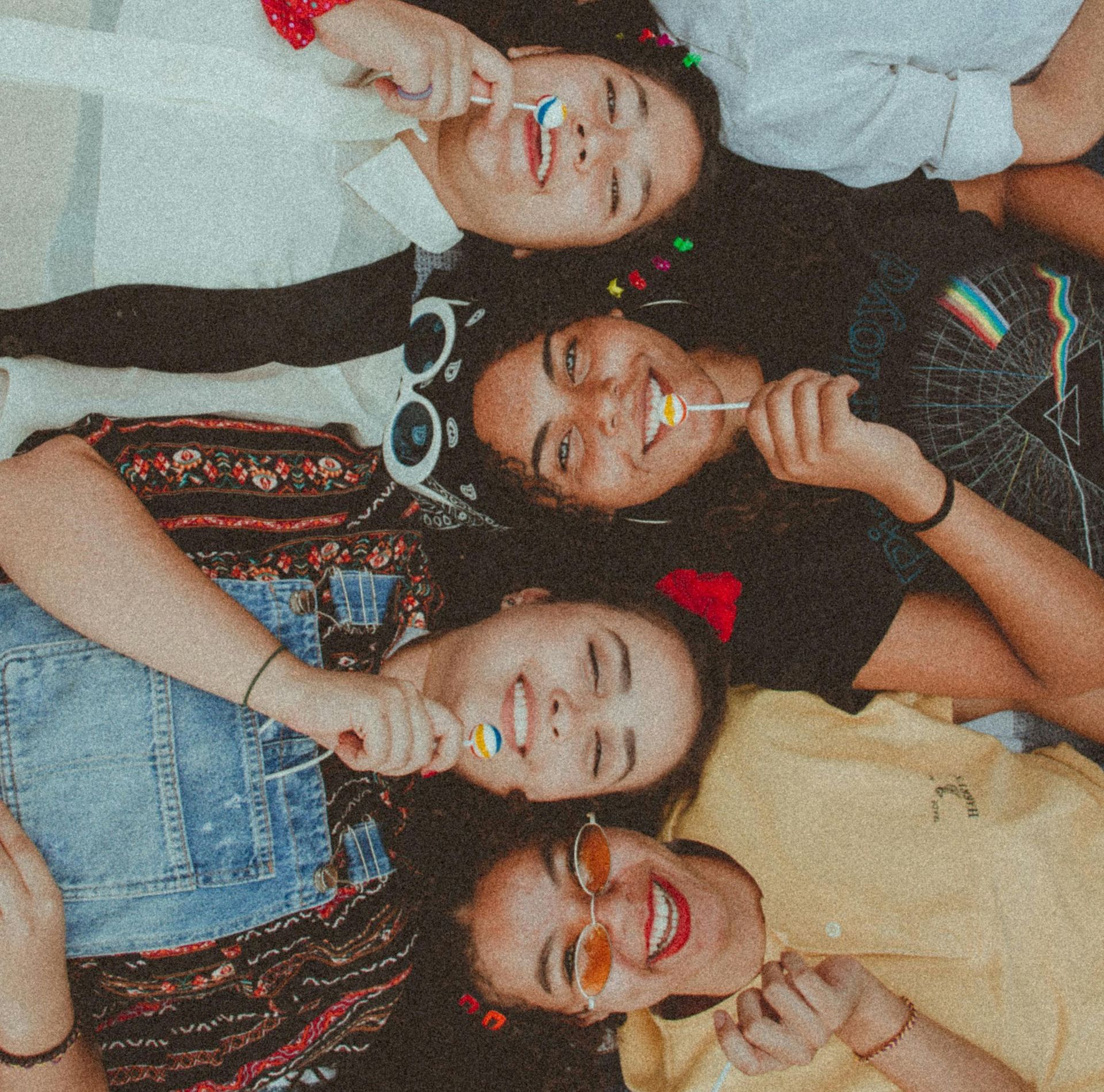 Four smiling people lying head-to-head, holding lollipops. Wearing casual clothes, sunglasses; outdoors.