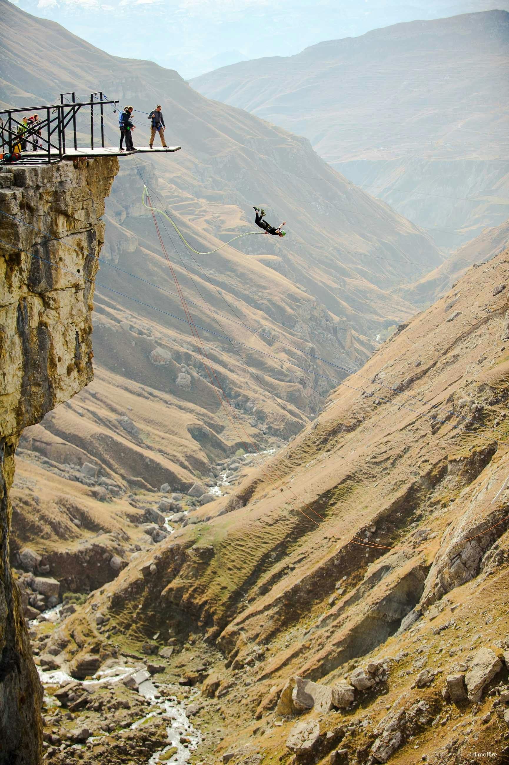 Person base jumping off cliff into canyon, two people watch from platform.