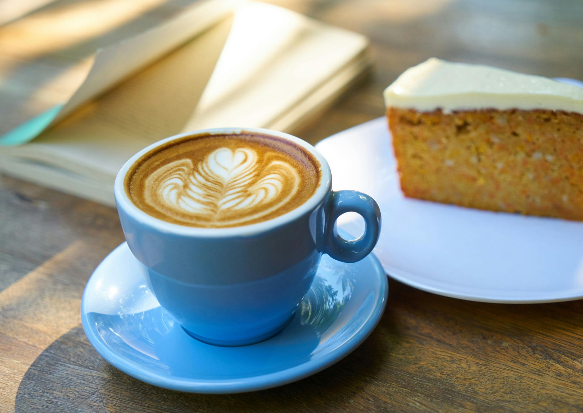 A cup of latte art on a saucer, a slice of carrot cake, and an open book on a wooden table.
